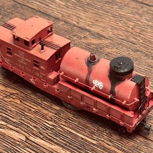 May include: A red model train caboose and tank car with black accents. The caboose has visible text and windows. The tank car has a black top and graffiti. The model sits on a wooden surface.