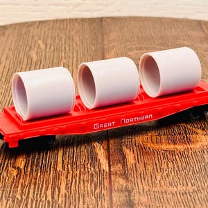 May include: A red toy train car with the words "Great Northern" printed on the side. The car is carrying three white cylindrical objects.