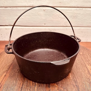 May include: A black cast iron Dutch oven with a curved metal handle. The pot has a rounded shape and sits on a wooden surface. The background is a white wooden wall.