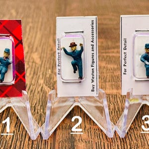 May include: Three miniature figures wearing blue uniforms and hats, displayed on clear plastic stands. Each stand has a white card with the text "For Perfect Detail" and "Buy Weston Figures and Accessories". The third stand also has the text "HEALDSBURG, CALIF."
