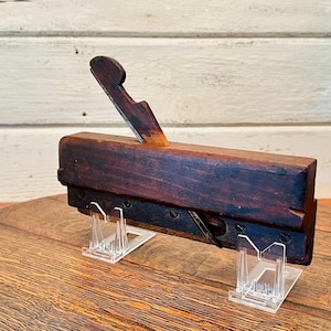 May include: Antique wooden hand plane with a metal blade, displayed on two clear plastic stands. The plane is dark brown with a worn finish.