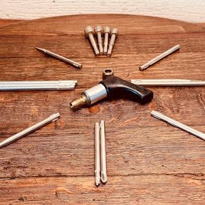 May include: A collection of metal tools, including a black and silver tool with a handle, several silver rods, and other metal pieces. The tools are arranged on a dark wooden surface.