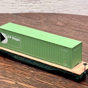 May include: A green CP Ships shipping container sits on a model train flatcar. The container has the CP Ships logo and is rectangular. The flatcar is green and brown, with the text A.T.&S.F. 90506.