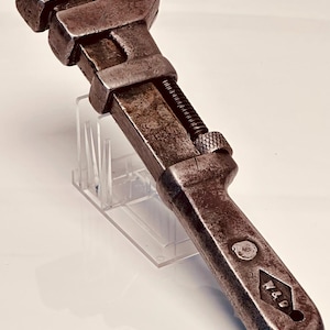 May include: An antique adjustable wrench with a weathered, metallic finish. The wrench features a movable jaw, a threaded adjustment mechanism, and the markings "U.P.R.R." and "W&B". The tool is displayed on a clear stand.