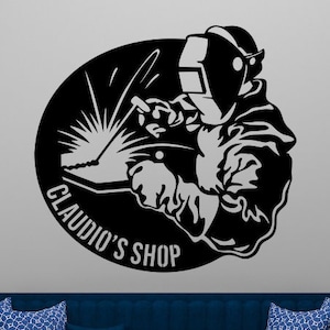 May include: A black and white silhouette of a welder with a welding torch, inside a circle with the text "Claudio's Shop". The image is on a gray wall behind a blue sofa with a coffee table and fruit bowl.