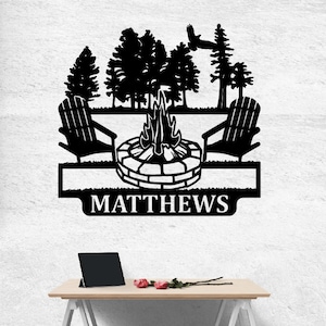 May include: Black metal wall art depicting a campfire scene with two Adirondack chairs, trees, and a bird in flight. The name "MATTHEWS" is shown below. A decorative piece for home or garden.
