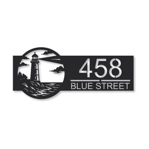 May include: Black metal address sign featuring a lighthouse scene. The lighthouse is on a rocky shore with clouds and water. The address numbers "458" and the words "BLUE STREET" are in silver.