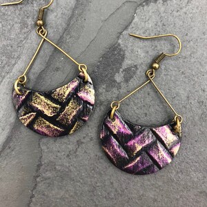 Può includere: Un paio di orecchini dorati e viola con un motivo intrecciato. Gli orecchini hanno la forma di una mezzaluna e pendono da un semplice filo dorato.