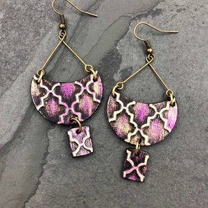 Può includere: Un paio di orecchini pendenti con un motivo geometrico. Gli orecchini sono in legno e hanno una finitura dorata. Il motivo è in tonalità di rosa e bianco.