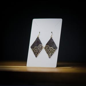 Puede incluir: Un par de pendientes de forma de diamante negro y dorado con un patrón geométrico. Los pendientes están colgados de una tarjeta de exhibición blanca.