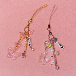 May include: Two decorative phone charms with butterfly and heart pendants. One charm is gold with pink accents, the other is silver with iridescent and white accents. Both charms have the letters "ΣΩΝ" on them.