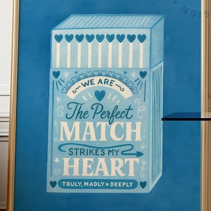 Pode incluir: Uma impressão artística emoldurada com um design de caixa de fósforos azul com o texto "The Perfect Match Strikes My Heart". O design da caixa de fósforos é adornado com corações e as palavras "We Are" e "Truly, Madly, Deeply". A obra de arte tem um fundo azul.