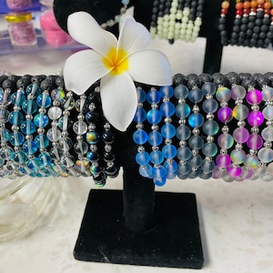 以下が含まれることがあります： 虹色のガラスビーズと黒い溶岩ビーズで作られた12本のブレスレットのセット。ブレスレットは、青から紫へのグラデーションで並んでいます。