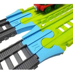 May include: A toy train track set featuring two parallel gray plastic tracks connected to bright lime green track sections by light blue plastic adapters. A small red and green toy train engine is visible on the green track section.