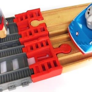 May include: A red and gray toy train on a gray track, connected to a wooden track with a blue toy train. The red train has a black window and a wooden front. The blue train has a face.