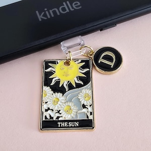 Puede incluir: Un dije dorado con borde negro que presenta un sol amarillo y margaritas blancas, con el texto "THE SUN". También hay un dije adicional con la letra "D". El dije está adjunto a un Kindle.