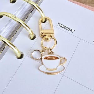Könnte beinhalten: Ein goldfarbener Schlüsselanhänger mit einem Kaffeetassen-Anhänger. Der Anhänger ist weiß mit braunem Kaffee und sitzt auf einer weißen Untertasse. Der Schlüsselanhänger ist an einer Planerseite mit "DONNERSTAG" befestigt.