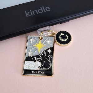 Puede incluir: Un dije dorado con un diseño de carta del tarot con un gato negro, estrellas y el texto "THE STAR". El dije está sujeto a un lector Kindle. También hay un dije circular con un diseño negro y dorado.