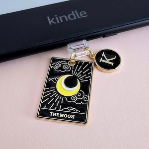 Puede incluir: Un charm de carta de tarot negro y dorado con un diseño de media luna amarilla y las palabras "THE MOON". También hay un charm dorado con la letra "K" adjunta. Los charms cuelgan de un clip transparente, cerca de un Kindle.