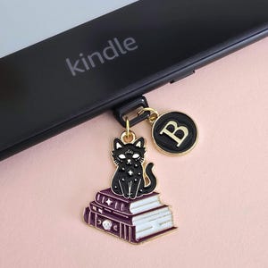 Op de afbeelding: Een zwarte Kindle e-reader met een bedel met een zwarte kat op boeken. De bedel heeft gouden details en een ronde hanger met de letter "B". Het woord "kindle" staat op de e-reader.