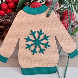 Peut inclure: Une décoration de Noël en papier beige en forme de pull avec un flocon de neige vert. L'ornement a une bordure verte et un trou en haut pour l'accrocher.