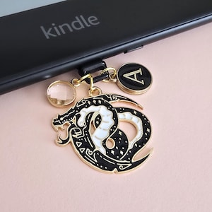 Puede incluir: Un lector de libros electrónicos Kindle negro con un dije decorativo. El dije presenta un diseño de dragón en blanco y negro, un dije con la inicial "A" de color dorado y una cuenta de cristal transparente. La palabra "kindle" está impresa en el lector.