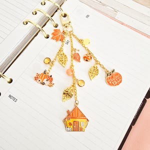 Pode incluir: Um pingente de agenda em metal dourado com vários elementos pendurados, incluindo folhas de esmalte laranja, abóboras, uma pequena casa e um pingente de abóbora redondo com o texto Hello Fall, preso a um fecho lagosta.