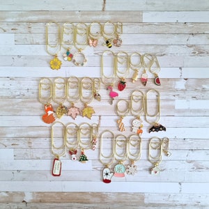 May include: A set of gold binder clips with various charms attached. The charms include a butterfly, a strawberry, a flamingo, a Christmas tree, a snowflake, and a fox. The clips are arranged in rows on a white wooden background.