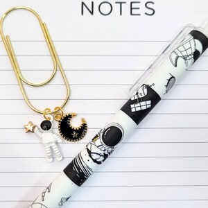 Puede incluir: Un clip de papel dorado con un dije de un astronauta y una luna creciente. El clip de papel está unido a un cuaderno con la palabra "NOTES" impresa en la parte superior. Un bolígrafo blanco y negro con un diseño de astronauta y temática espacial está sobre el cuaderno.
