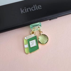 Può includere: Un ciondolo a forma di libro verde e oro con un pendente a forma di gemma verde attaccato a un Kindle. Il ciondolo a forma di libro ha dettagli bianchi ed è attaccato a una clip verde trasparente. Il Kindle è nero con la scritta "Kindle".