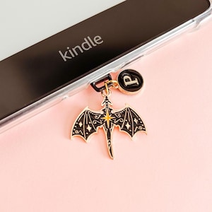Puede incluir: Un dije de dragón negro y dorado con un diseño de estrella cuelga de una funda de teléfono junto a un círculo negro con la letra "P". La palabra "kindle" es visible en la pantalla del teléfono. El dije está sobre un fondo rosa.