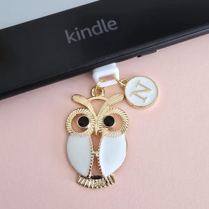 Puede incluir: Un dije de búho dorado con ojos negros y esmalte blanco, colgando de un Kindle. También se adjunta un pequeño dije redondo con la letra "N". El Kindle es negro y el fondo es rosa claro.
