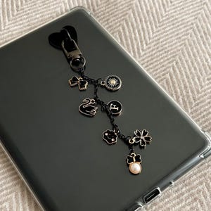 May include: Black and gold charm keychain with a heart-shaped clasp. The charms include a bow, sunburst, letter "F", swan, flower, and a pearl. The keychain is resting on a black surface.