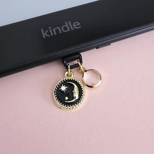 Puede incluir: Lector de libros electrónicos Kindle negro con un dije con un diseño de luna y estrella negro y dorado, y un dije redondo transparente. La palabra "kindle" está impresa en el dispositivo. El fondo es rosa pálido.