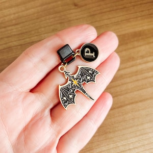 May include: A black and gold dragon charm with a yellow star accent, attached to a black clasp and a circular charm with the letter "P". The dragon has detailed wing and body designs. The charms are likely for a keychain or zipper pull.
