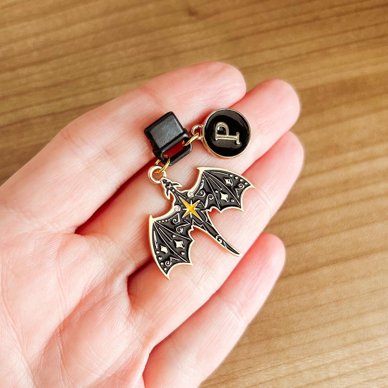 May include: A black and gold dragon charm with a yellow star accent, attached to a black clasp and a circular charm with the letter "P". The dragon has detailed wing and body designs. The charms are likely for a keychain or zipper pull.