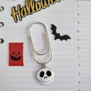 Könnte beinhalten: Eine silberne Büroklammer mit einem Jack Skellington-Anhänger, einer schwarzen Fledermaus und einem roten Halloween-Süßigkeitentüten-Aufkleber auf einem linierten Papierhintergrund. Das Wort "Halloween" steht in Gold oben. Artikel für Planer oder Scrapbooking.