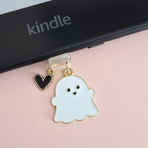 Puede incluir: Un charm de fantasma blanco con contorno dorado y un charm de corazón negro colgando de un clip de plástico transparente. La palabra "kindle" es visible en un dispositivo electrónico negro.