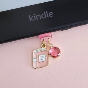 Könnte beinhalten: Ein rosa-goldener Buchanhänger und ein rosafarbener Edelstein hängen an einem durchsichtigen rosa Clip, der an einem Kindle befestigt ist. Das Wort "kindle" ist auf dem Gerät aufgedruckt. Der Hintergrund ist ein zartes Rosa.