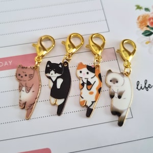 May include: Five gold cat charms with different colored cat designs. The charms are attached to gold lobster clasps. The charms are on a white and gold lined background.