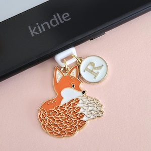 Puede incluir: Un charm decorativo con un zorro naranja estilizado, detalles blancos y dorados, colgado de un cierre blanco. También hay un charm circular blanco con borde dorado con la letra "R". Cerca de un Kindle negro.