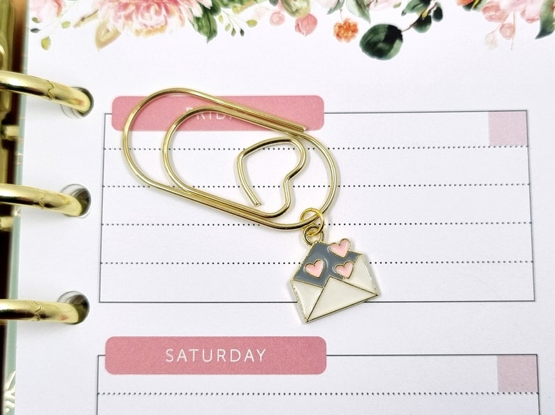 K&ouml;nnte beinhalten: Ein goldener B&uuml;roklammer mit einem kleinen Briefumschlag-Anh&auml;nger mit rosa Herzen. Der Briefumschlag-Anh&auml;nger ist mit einem goldenen Ring an der B&uuml;roklammer befestigt. Die B&uuml;roklammer befindet sich auf einer wei&szlig;en Seite mit rosa Linien und dem Wort "SAMSTAG" auf einem rosa Balken.