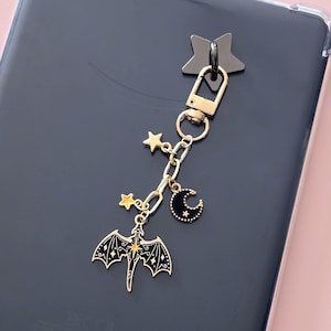 Puede incluir: Un llavero dorado con un cierre en forma de estrella negra. La cadena tiene colgantes de un dragón, estrellas y una luna creciente. El colgante del dragón tiene alas negras y un centro amarillo. El colgante de la luna es negro con estrellas blancas.