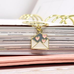 K&ouml;nnte beinhalten: Ein goldfarbener Briefumschlag-Anh&auml;nger mit einem wei&szlig;en und grauen Emaille-Design und drei rosa Herzen. Der Anh&auml;nger ist an einem goldfarbenen Ring befestigt.