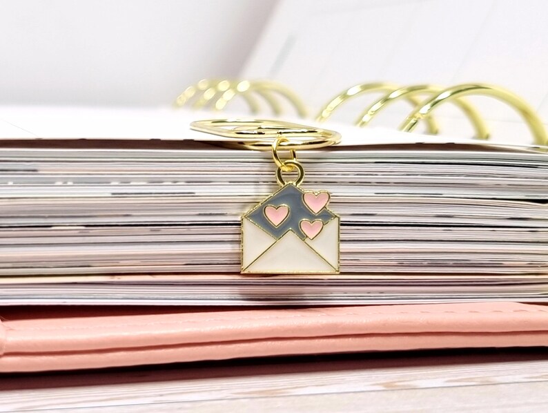 K&ouml;nnte beinhalten: Ein goldfarbener Briefumschlag-Anh&auml;nger mit einem wei&szlig;en und grauen Emaille-Design und drei rosa Herzen. Der Anh&auml;nger ist an einem goldfarbenen Ring befestigt.