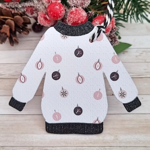 Può includere: Etichetta regalo a forma di maglione natalizio bianco con un cordoncino in iuta nero e bianco. Il maglione è decorato con ornamenti rosa e marroni.