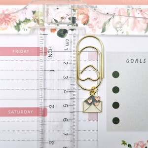 K&ouml;nnte beinhalten: Ein goldfarbener B&uuml;roklammer mit einem kleinen Briefumschlag-Anh&auml;nger mit drei rosa Herzen. Der Anh&auml;nger ist mit einer goldenen Kette an der B&uuml;roklammer befestigt. Die B&uuml;roklammer befindet sich auf einer wei&szlig;en Planerseite mit den W&ouml;rtern "FRIDAY" und "SATURDAY". Die Planerseite hat auch einen Abschnitt mit der Bezeichnung "GOALS" mit drei schwarzen Punkten.