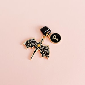 May include: A black and gold dragon charm with a star design and a round charm with the letter "P" on a pink background. The dragon has detailed wings and is attached to a black clasp.