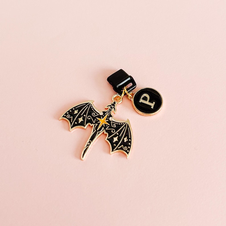 May include: A black and gold dragon charm with a star design and a round charm with the letter "P" on a pink background. The dragon has detailed wings and is attached to a black clasp.