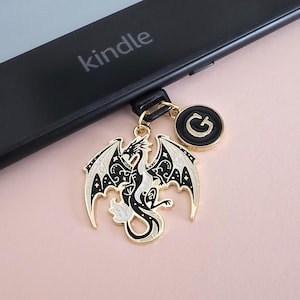Puede incluir: Un charm de dragón negro y dorado con vientre y alas blancas, colgado de un dispositivo Kindle. El charm tiene un colgante circular con la letra "G". El Kindle es negro y tiene la palabra "kindle" impresa.